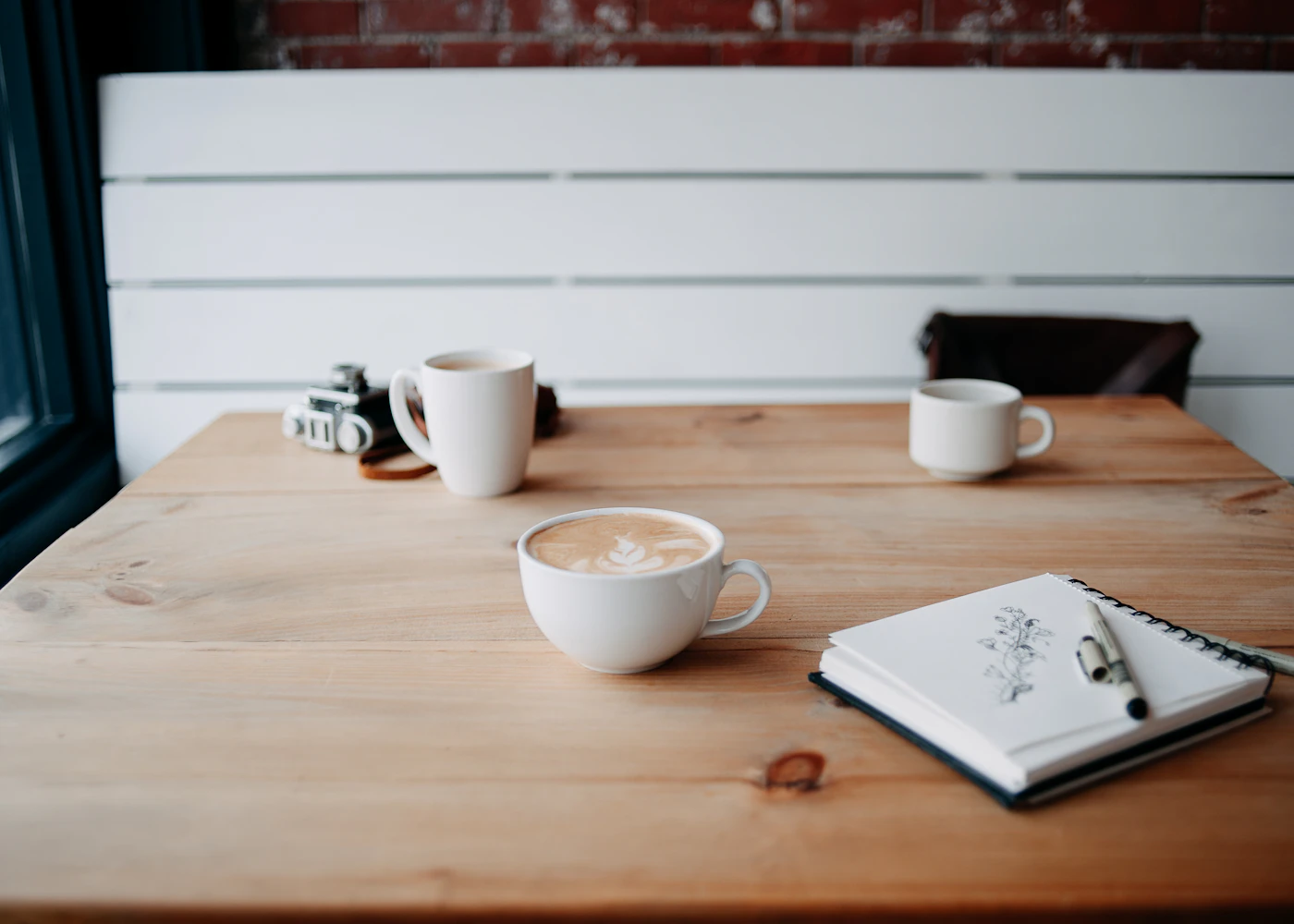 Coffee cups and an open sketchbook on a wooden cafe table by a window